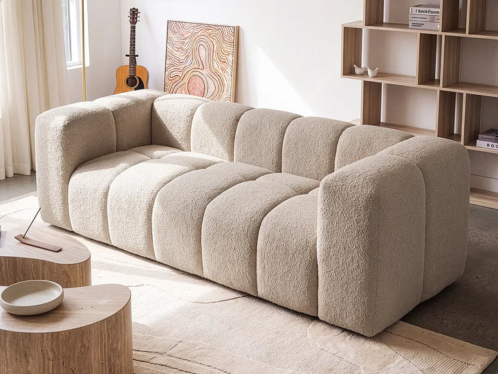 A modern beige bouclé sofa with a segmented, bubble-like design, styled in a bright, minimalist living room with wooden tables, a bookshelf, and an acoustic guitar in the background.