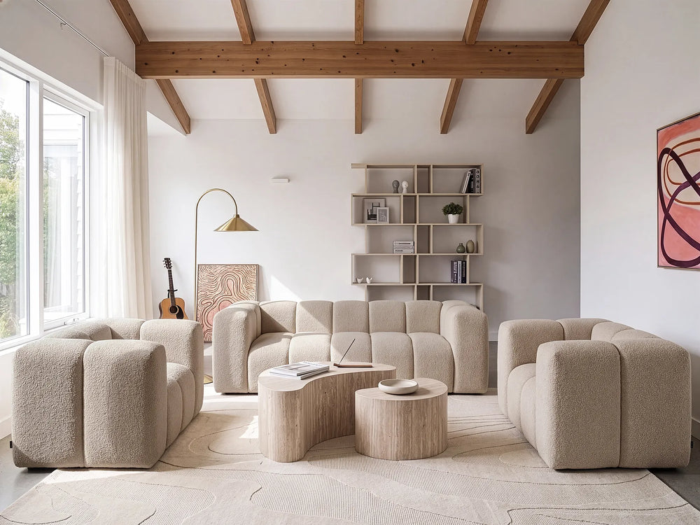 A modern living room featuring a modular beige bouclé sofa, two matching armchairs, two curved light wood coffee tables, a floor lamp, and a built-in shelving unit against a white wall with exposed wooden ceiling beams.
