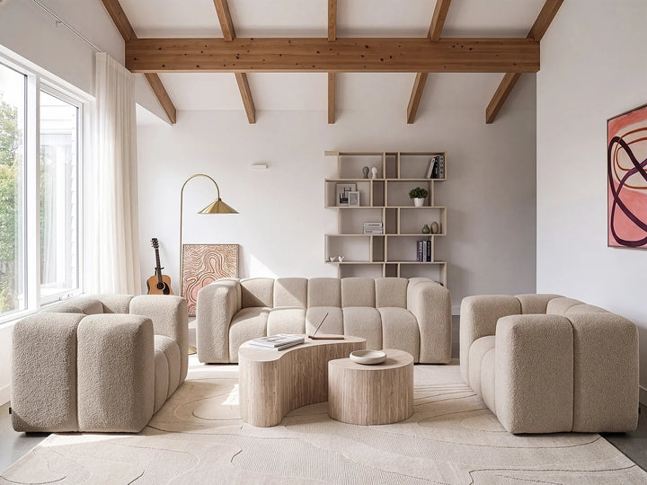 A modern living room featuring a modular beige bouclé sofa, two matching armchairs, two curved light wood coffee tables, a floor lamp, and a built-in shelving unit against a white wall with exposed wooden ceiling beams.