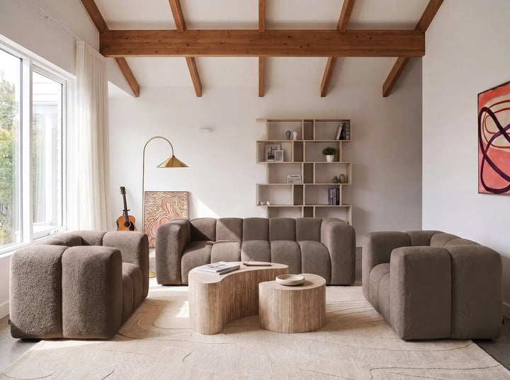 A modern living room featuring a modular, chunky boucle sofa set in taupe. The set includes a three-seater sofa, two matching armchairs, and two organic-shaped light wood coffee tables.