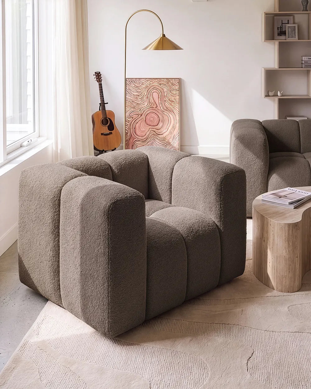 A modern, chunky upholstered armchair in a textured taupe fabric, styled in a bright, contemporary living room next to a wooden side table and an acoustic guitar leaning against the wall.