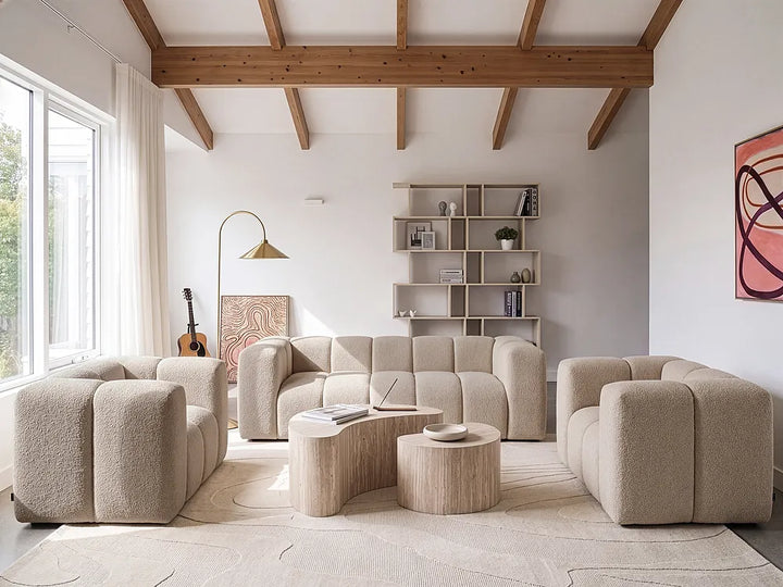 A modern living room featuring a modular, textured beige boucle sofa set with matching armchairs, two curved wooden coffee tables, a tall floor lamp, and a light wood shelving unit against a white wall with exposed wooden ceiling beams.