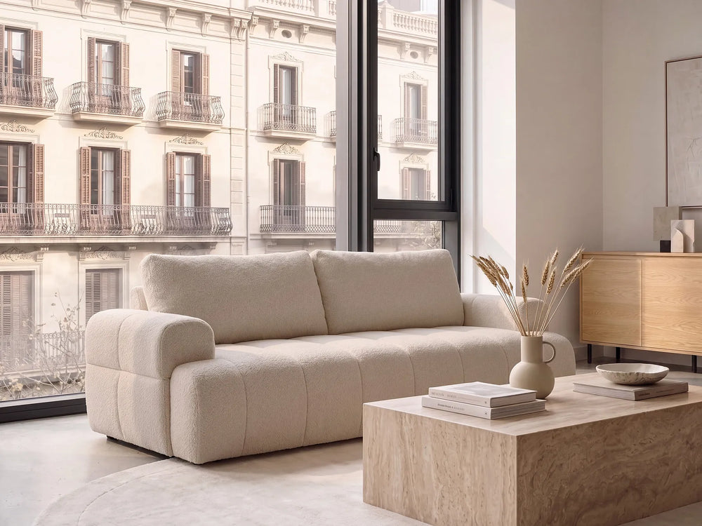 A modern beige bouclé three-seater sofa in a bright, contemporary living room with a large window overlooking classic apartment buildings, a wooden coffee table, and a light wood cabinet.