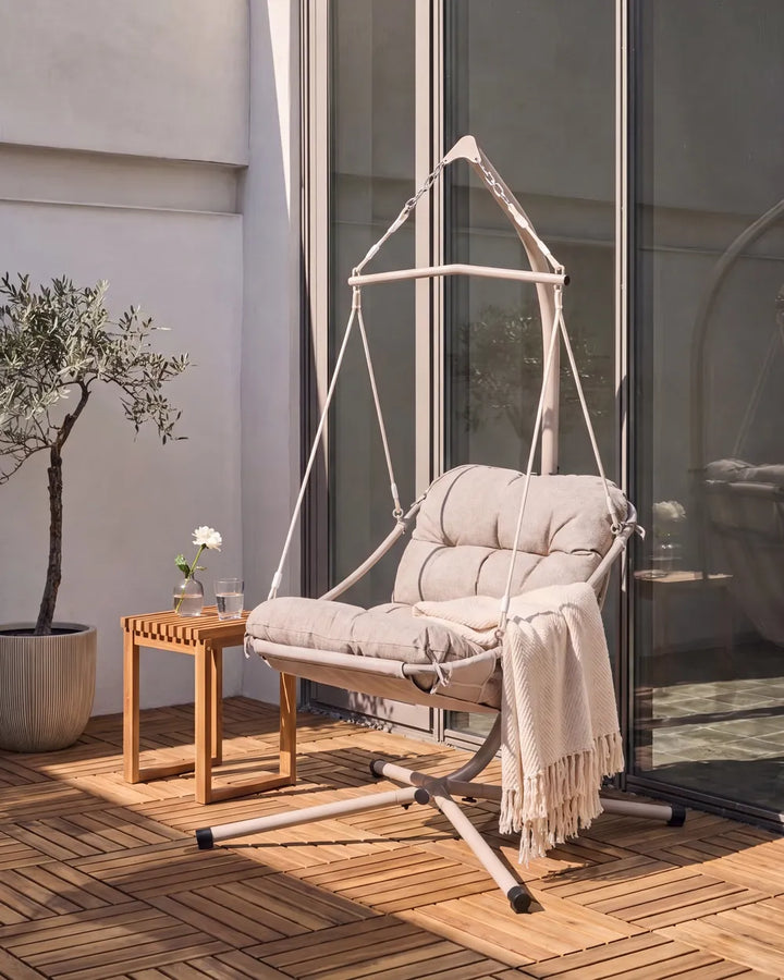 A cream-colored fabric hanging swing chair with a matching cushioned seat and backrest, styled on a wooden deck next to a small wooden side table with a throw blanket draped over it.