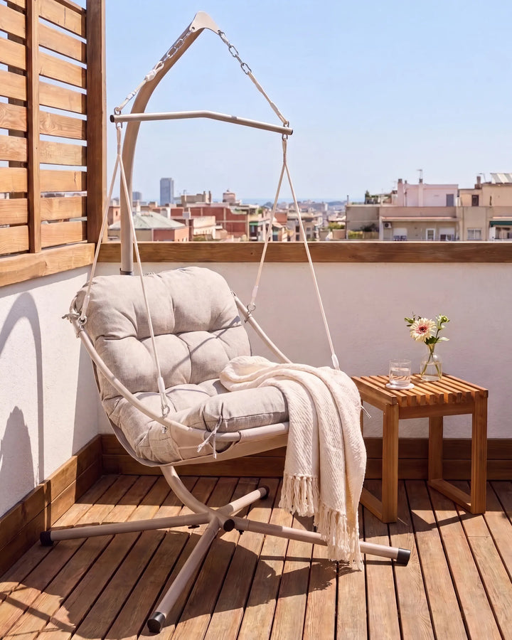 A beige fabric hanging hammock chair with a matching cushioned seat and a draped throw blanket, set on a wooden deck balcony with a small wooden side table and a city view in the background.