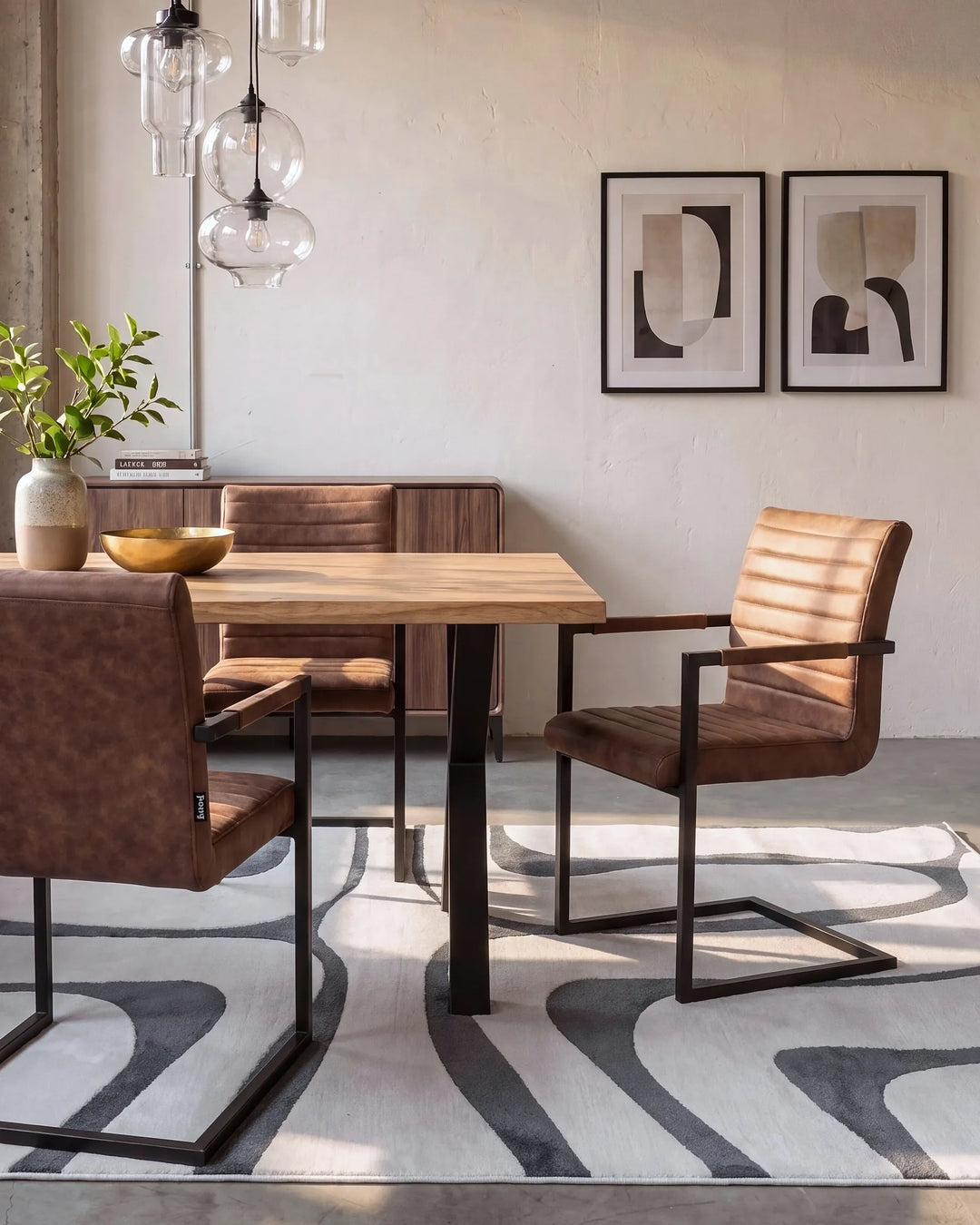 A dining room scene with a rectangular wooden table and three brown upholstered chairs with black metal frames, set on a patterned rug against a light textured wall with framed abstract art and hanging glass pendant lights.