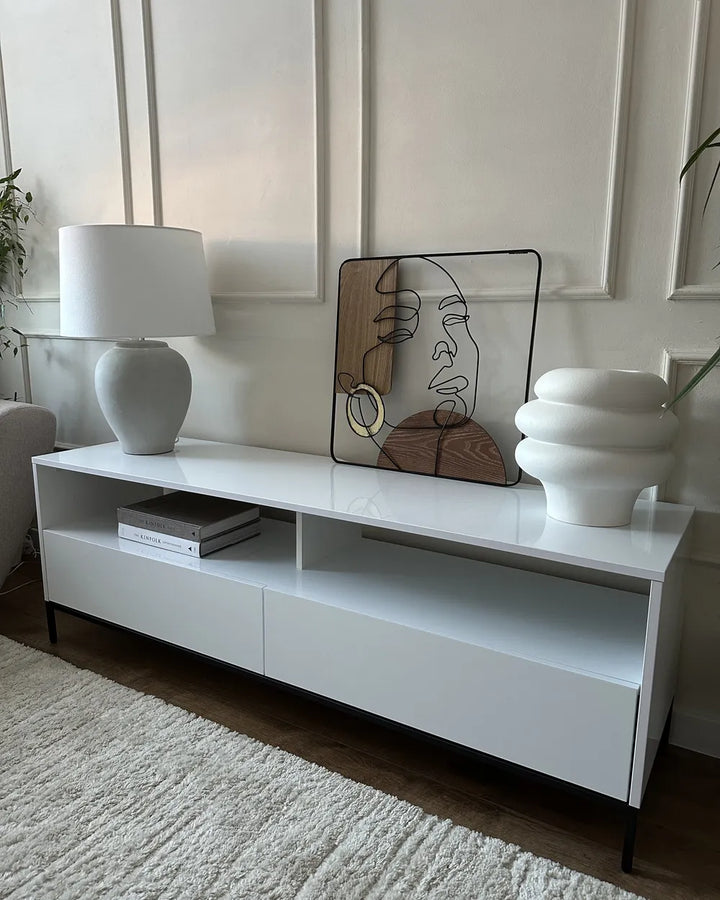 A modern white low-profile media console with open shelving and black legs, styled in a living room setting with a white table lamp, a framed abstract face print, and a white sculptural vase.