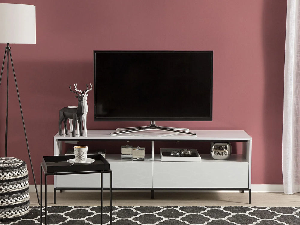 A modern white TV stand with open shelving and two lower drawers, styled in a living room against a dusty rose wall with a black floor lamp, patterned rug, and side table.