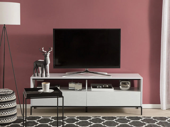 A modern white TV stand with open shelving and two lower drawers, styled in a living room against a dusty rose wall with a black floor lamp, patterned rug, and side table.