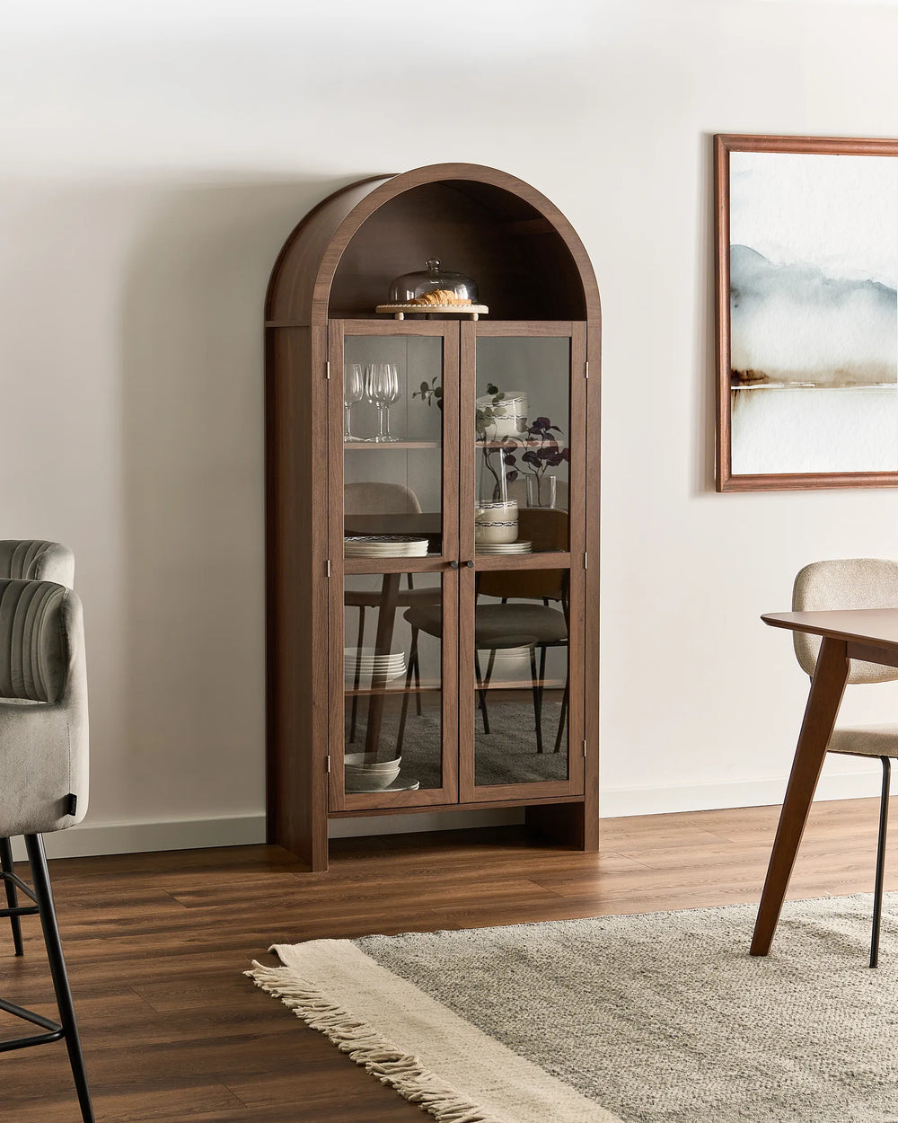 An arched wooden display cabinet with glass doors stands in a styled dining room setting with wood floors, a rug, and dining furniture.