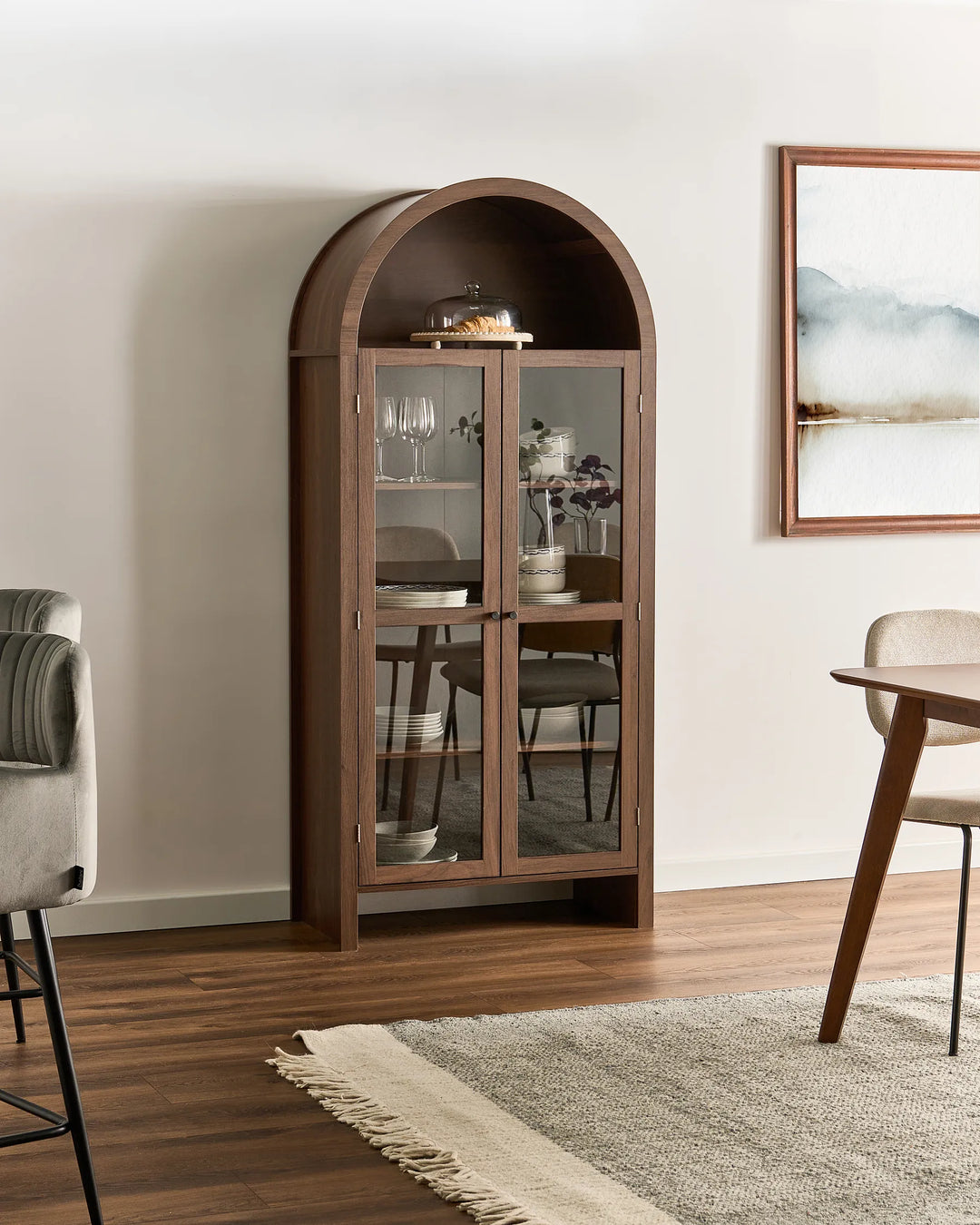 An arched wooden display cabinet with glass doors stands in a styled dining room setting with wood floors, a rug, and dining furniture.