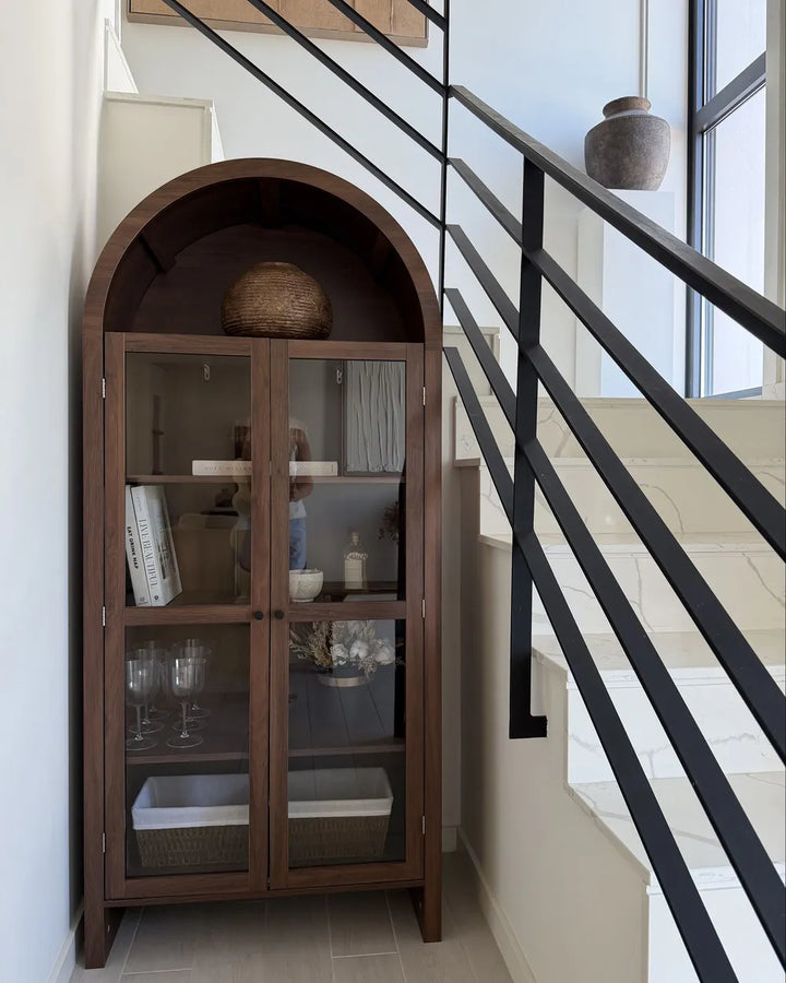 A tall arched wooden display cabinet with glass doors stands beside a staircase in a bright interior. The cabinet has a dark brown wood finish, glass panels, and open shelving with decorative items including a woven basket and books.