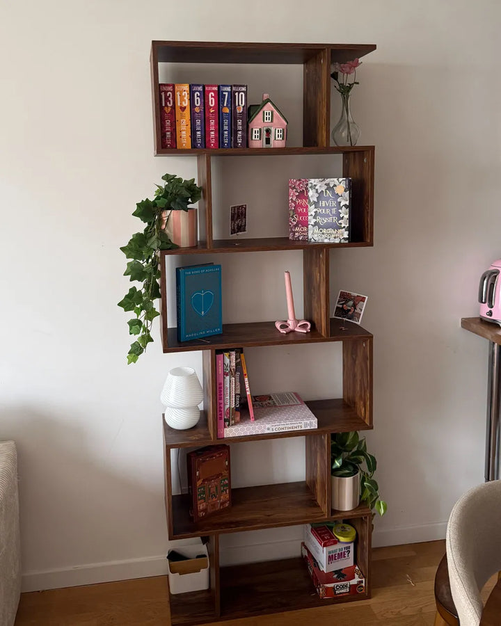 A tall, rustic wooden bookshelf with multiple open shelves styled with books, small plants, a pink candle, and decorative items, set against a plain white wall in a home interior.