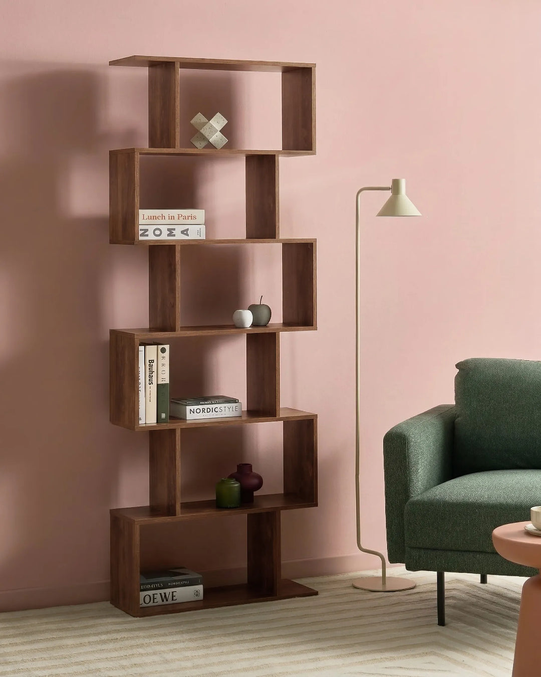 A modern, multi-tier wooden bookshelf with an open, geometric design in a warm medium-brown finish, styled in a living room setting against a dusty pink wall next to a green armchair and a floor lamp.
