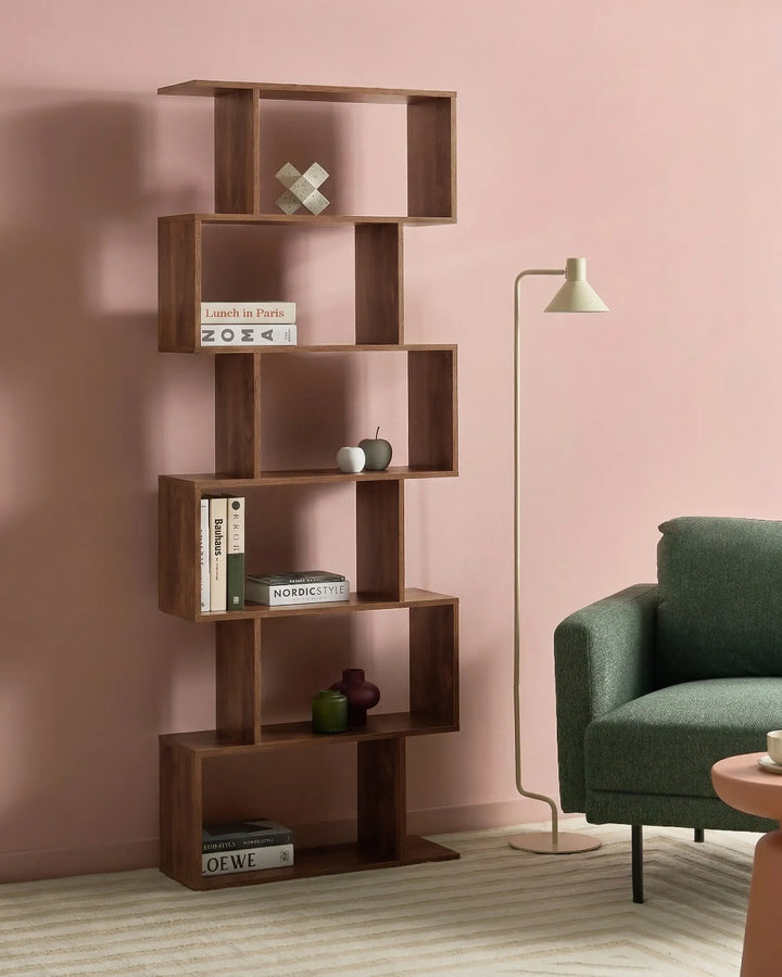 A modern, multi-tier wooden bookshelf with an open, geometric design in a warm medium-brown finish, styled in a living room setting against a dusty pink wall next to a green armchair and a floor lamp.