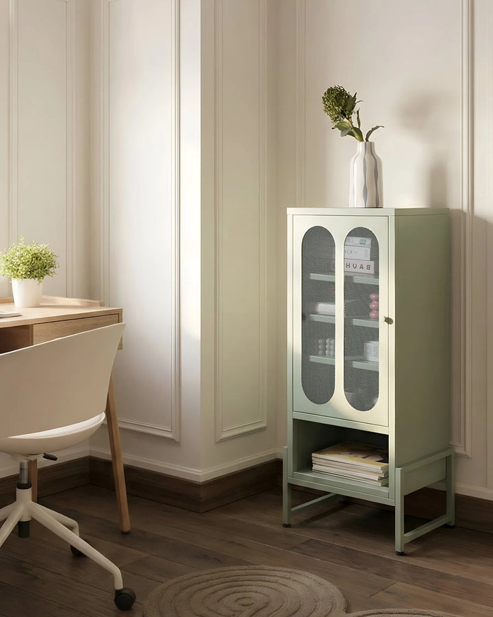 A sage green metal storage cabinet with glass doors and a lower shelf, styled in a bright home office next to a desk and chair.
