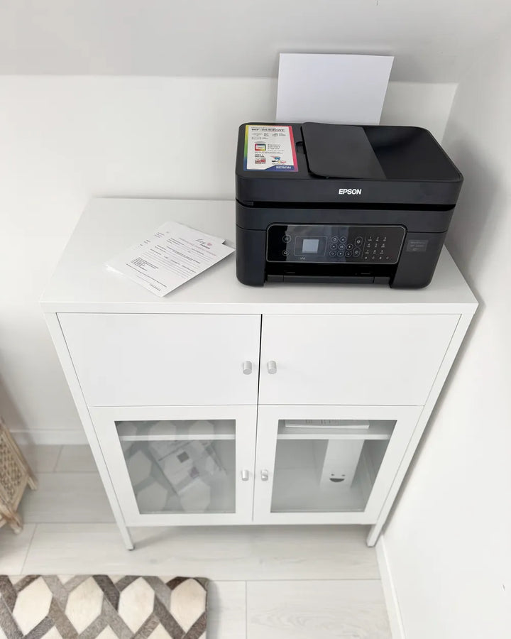 An Epson all-in-one wireless printer in black sits on top of a white two-door cabinet with glass doors, in a home office or study setting.