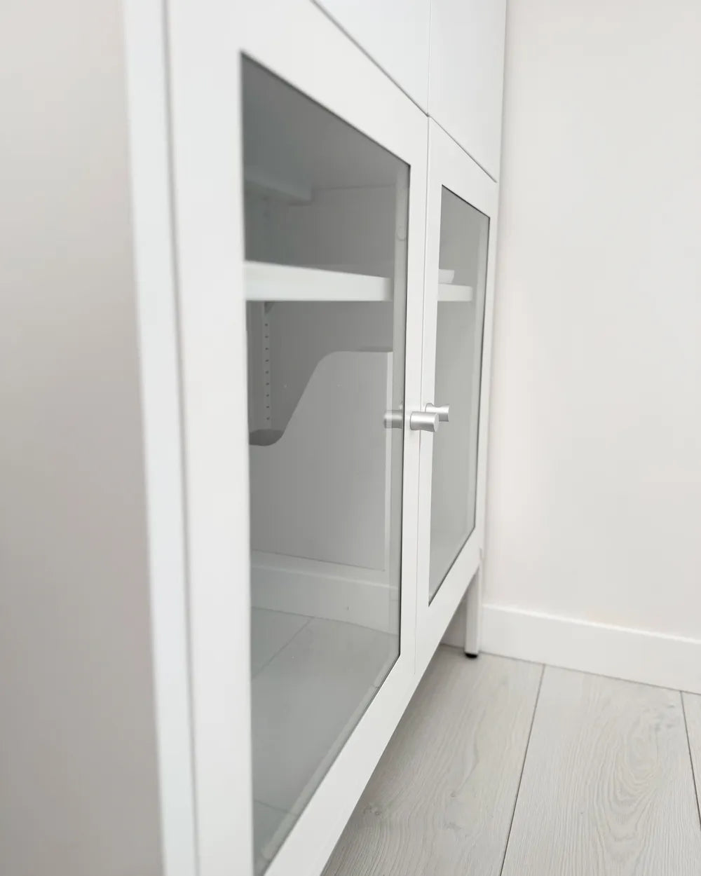 White storage cabinet with two glass doors and silver handles, shown in a minimalist room with light gray wood floors and walls.