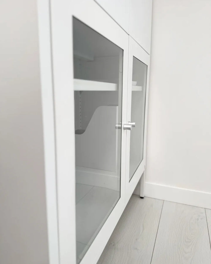 White storage cabinet with two glass doors and silver handles, shown in a minimalist room with light gray wood floors and walls.