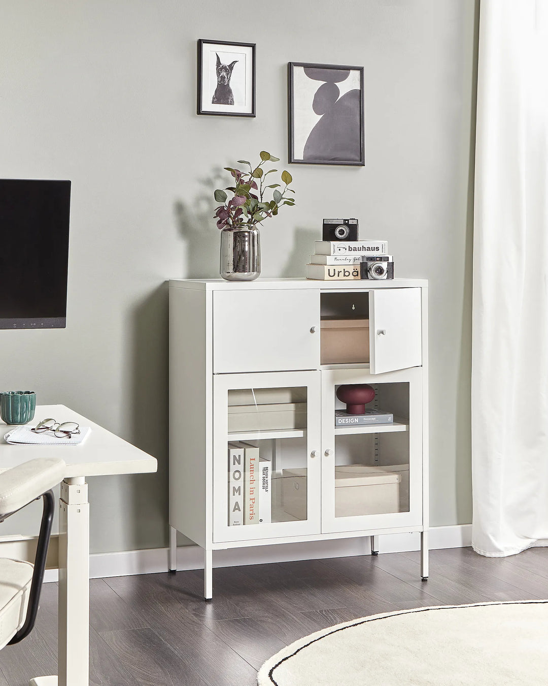 White storage cabinet with glass doors in a room with a desk and framed pictures on the wall.