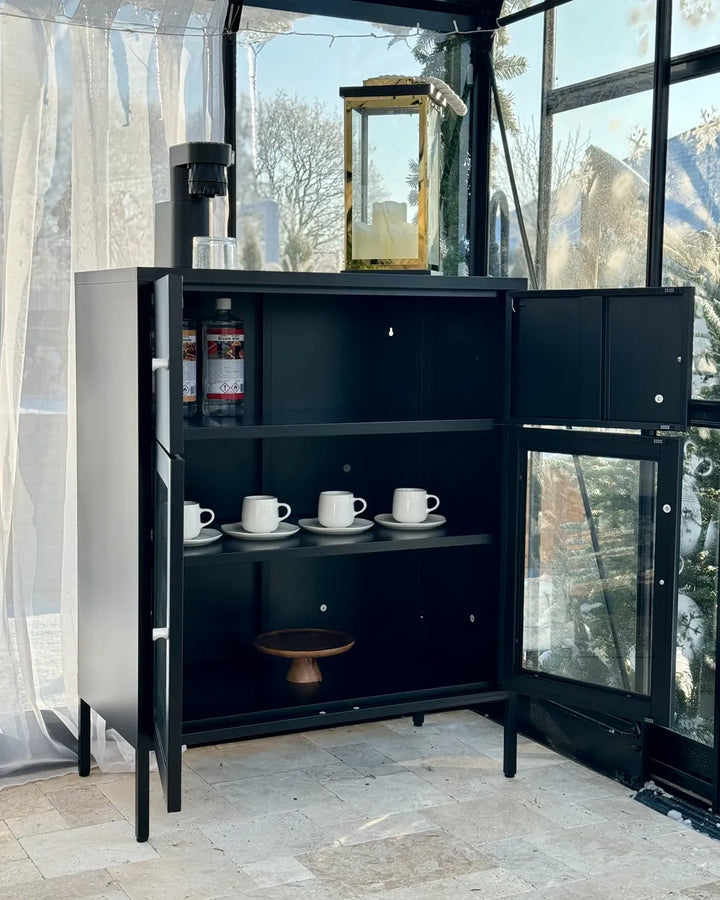 A tall black metal beverage station cabinet with open doors, shown in a glass-walled outdoor shelter. The cabinet has shelves holding white coffee mugs, bottles, and a small wooden tray, with a coffee maker and lantern on top.