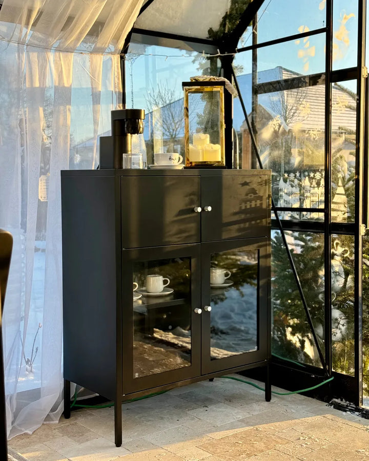A tall black cabinet with two upper doors and two lower glass doors, styled with coffee equipment and a lantern on top, set inside a glass-enclosed outdoor space.