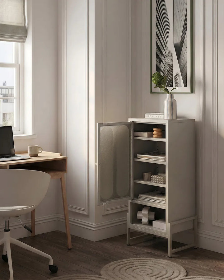 A tall, narrow light gray storage cabinet with an open door, shown in a styled home office setting next to a wooden desk and white chair.