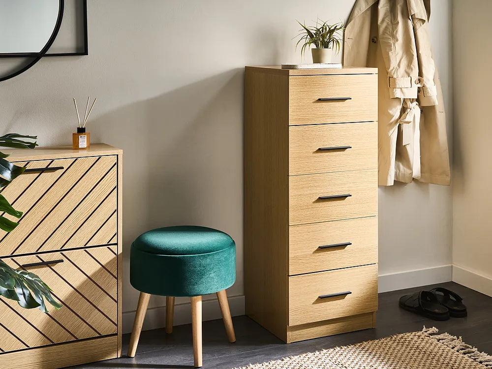 A tall light wood five-drawer chest in a styled room setting next to a green velvet stool, with other home decor items like a coat rack, mirror, and rug visible.