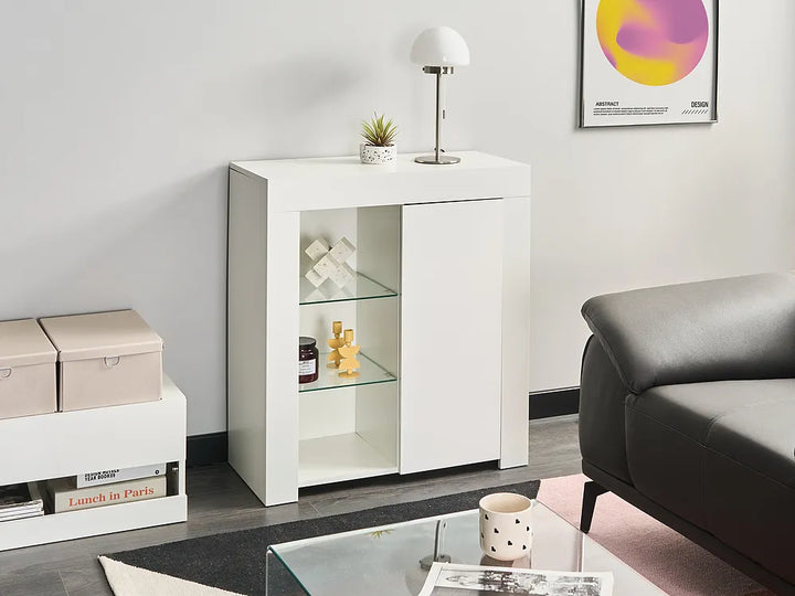 A modern white sideboard cabinet with two glass-front shelves and a right-hand door, styled in a minimalist living room next to a gray leather sofa.