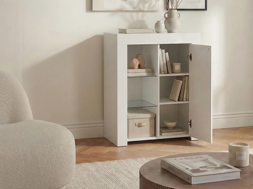 A white three-compartment side cabinet with one door open, styled in a bright living room next to a beige armchair and round coffee table.
