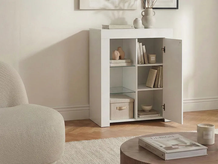 A white three-compartment side cabinet with one door open, styled in a bright living room next to a beige armchair and round coffee table.