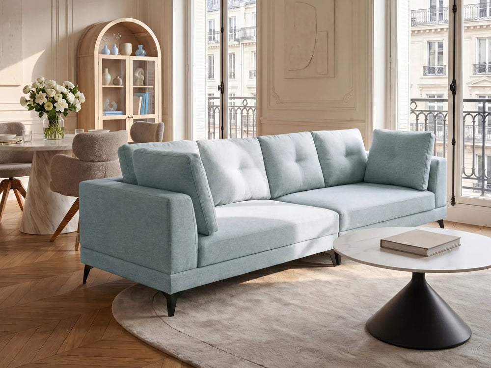 A light blue upholstered three-seat sofa with tufted back cushions, shown in a bright, elegant living room with wood floors, a round coffee table, and large windows.