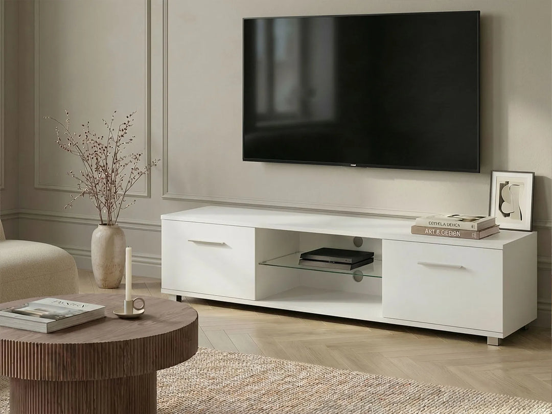 A modern white TV stand console with a glass shelf, shown in a styled living room setting with a wall-mounted television, a round wooden coffee table, and neutral decor.