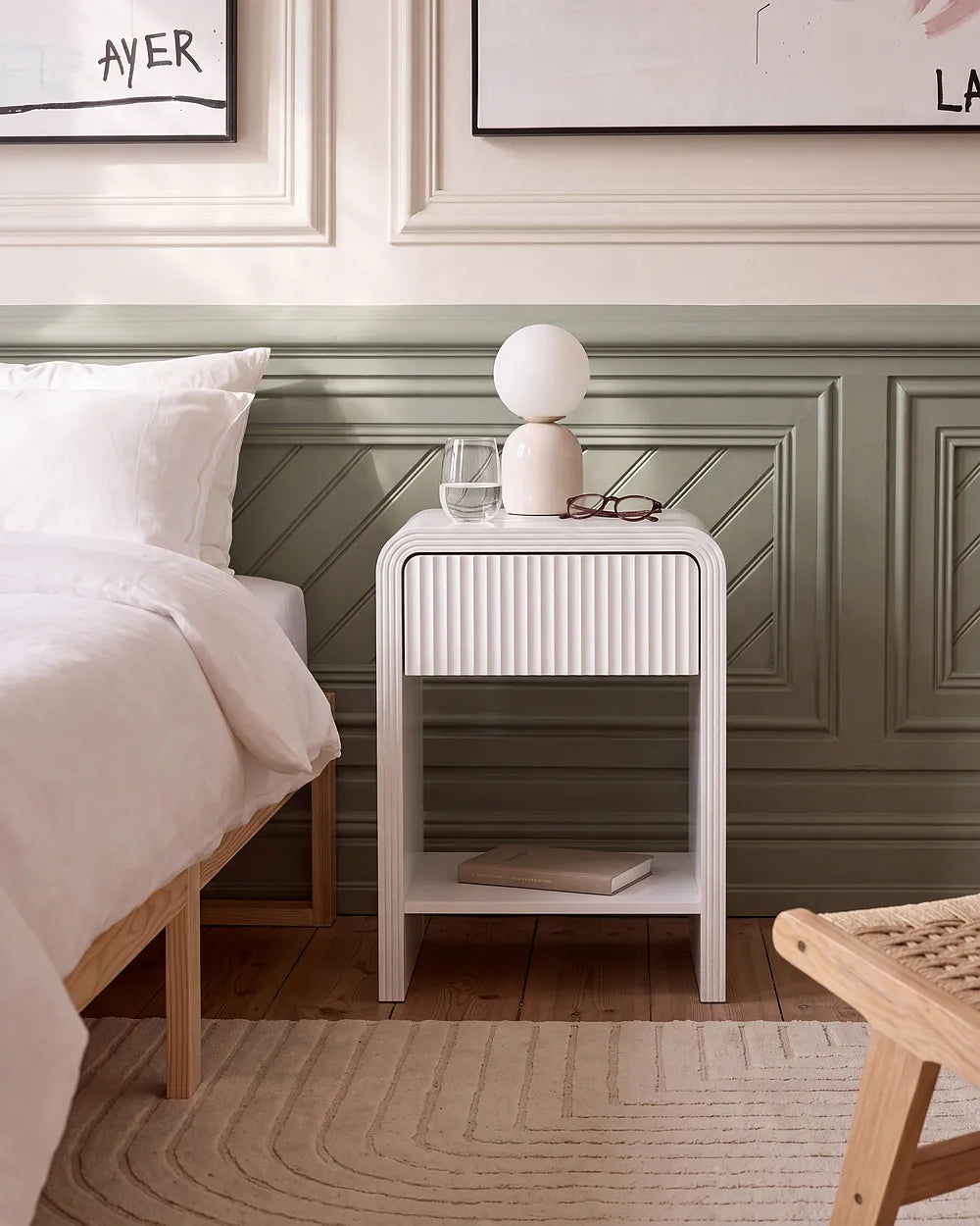 A modern white nightstand with a ribbed front and open lower shelf, styled in a bedroom next to a bed with white linens. The nightstand holds a spherical white lamp, a glass, and a pair of eyeglasses.