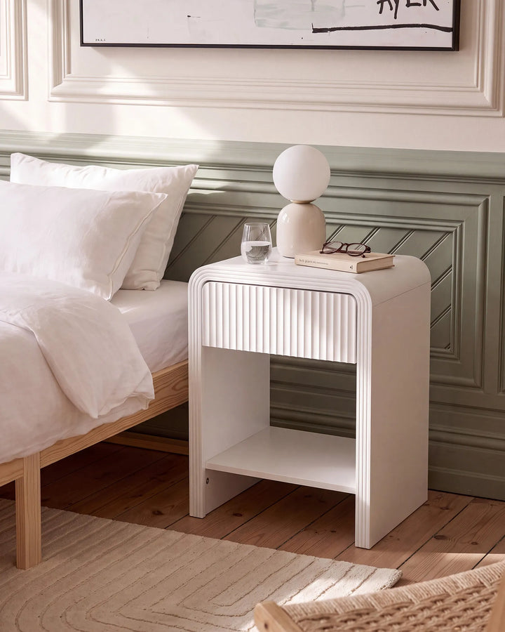 A modern white bedside table with a ribbed front and open lower shelf, styled next to a bed in a bright bedroom. The table holds a spherical lamp, a glass, and a book.