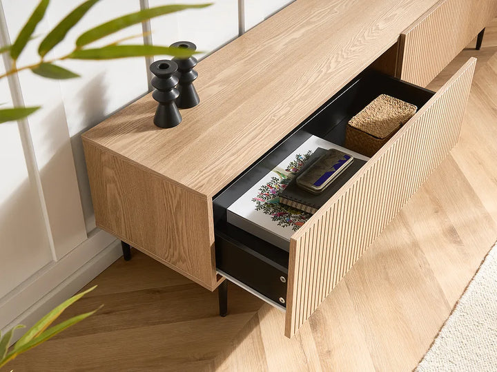 A modern light oak wood media console with a ribbed front drawer, shown with one drawer open to reveal storage. The console is styled in a bright room with light wood floors, white paneled walls, and a plant in the foreground.