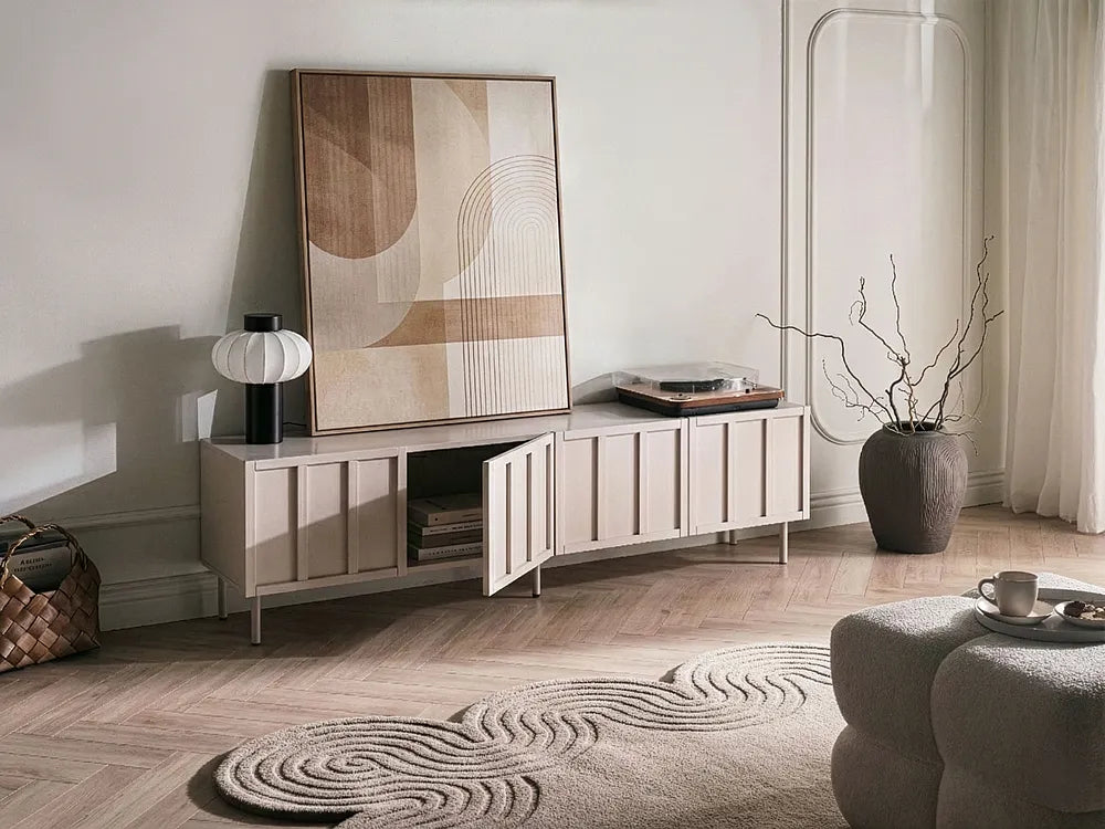 A modern light beige wooden media console with an open center cabinet, styled in a minimalist living room next to a large abstract art print, a black table lamp, and a gray textured vase with dried branches.