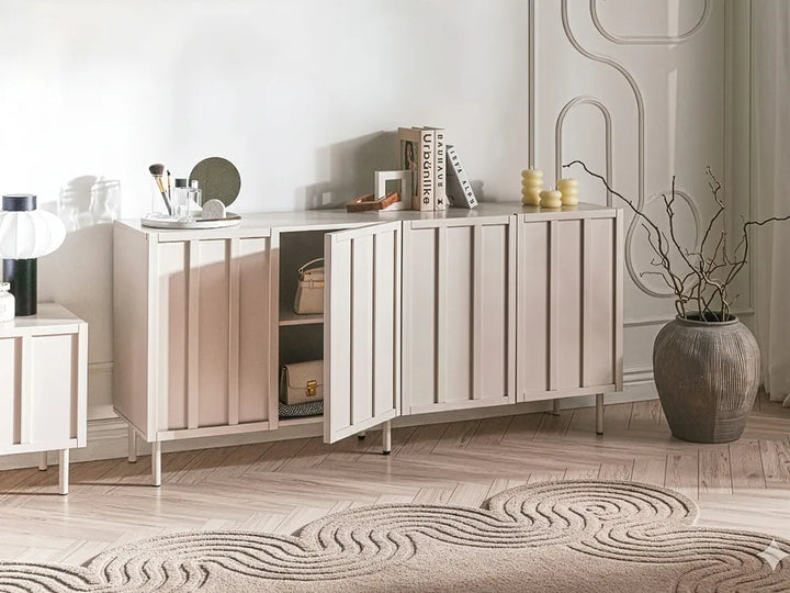 A light beige sideboard cabinet with vertical panel doors and a hinged open door, styled in a bright, modern room with a patterned rug, decorative books, and a large ceramic vase.
