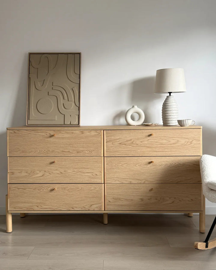 A light oak wood six-drawer dresser in a minimalist room setting, styled with a framed abstract print, a textured white lamp, and a small decorative object on top.