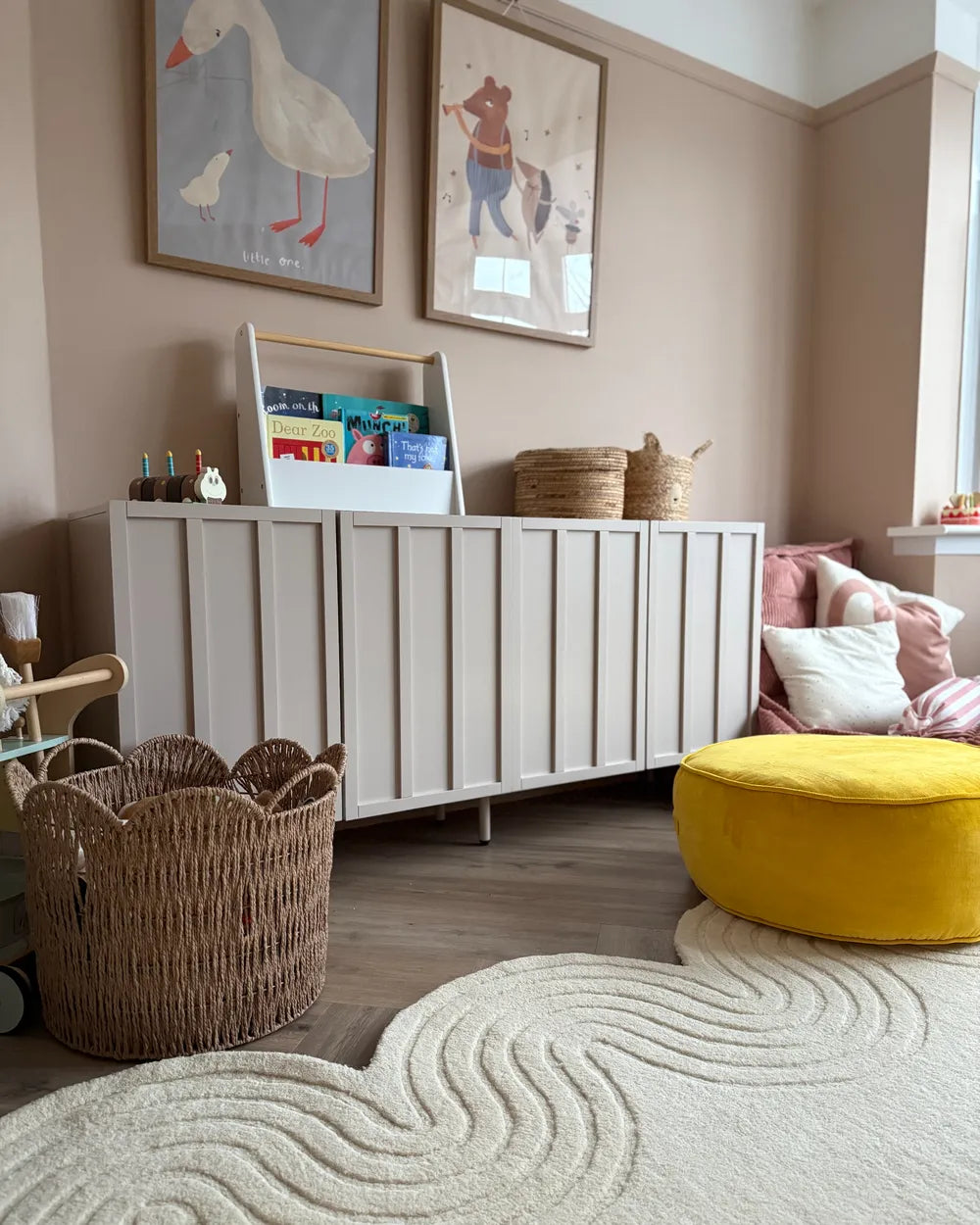 A styled children's room corner featuring a white slatted side cabinet with a bookshelf insert, a yellow velvet pouf, a cream wavy rug, woven baskets, and framed wall art against beige walls.