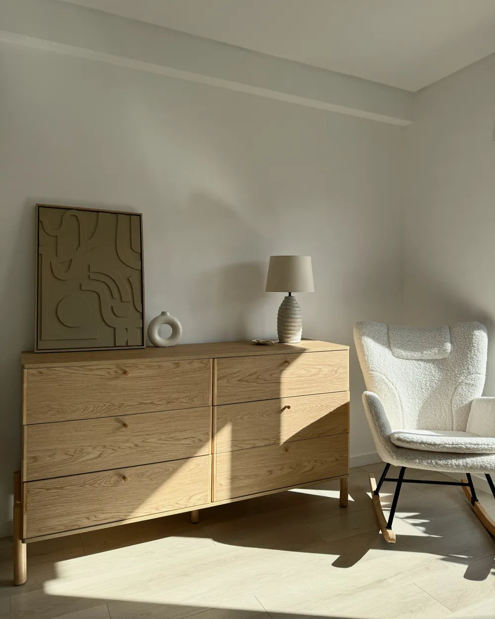 A light oak wood six-drawer dresser in a modern, sunlit room next to a white textured rocking chair and a small table lamp.