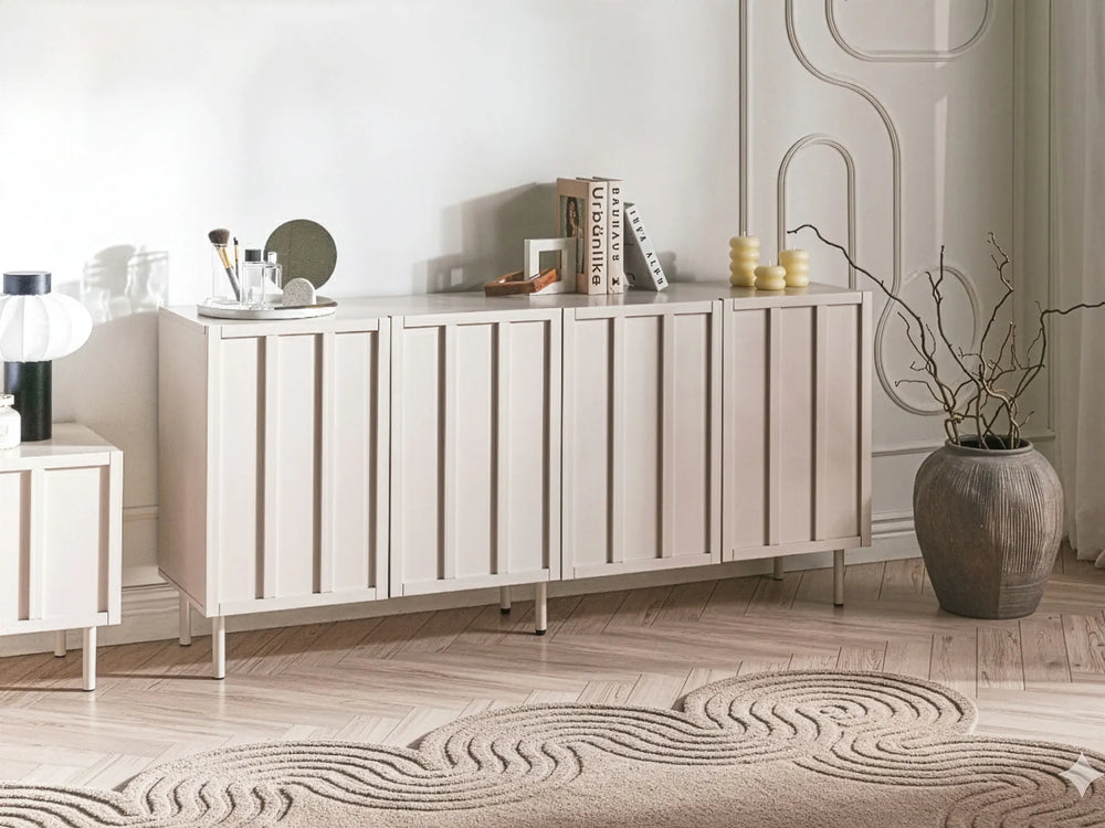 A modern light beige sideboard cabinet with vertical slat doors, styled in a minimalist room with a patterned rug, a large ceramic vase, and decorative objects on top.