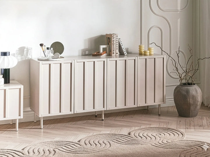 A modern light beige sideboard cabinet with vertical slat doors, styled in a minimalist room with a patterned rug, a large ceramic vase, and decorative objects on top.