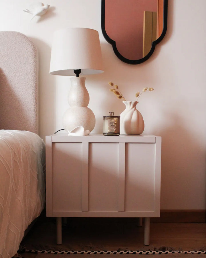 A modern white nightstand in a bedroom setting, styled with a matching white table lamp, a small candle, a ceramic vase with dried stems, and a pink wall mirror above.