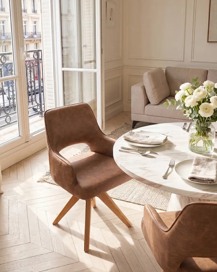 A modern brown upholstered dining chair with a curved back and light wood legs, shown in a bright, sunlit Parisian-style dining room next to a round white marble table with plates and a vase of white flowers.