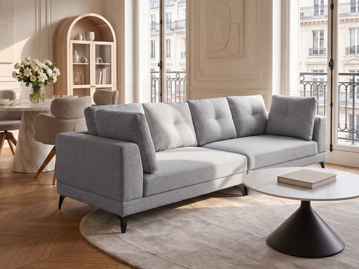 A modern light gray fabric sofa with tufted back cushions and tapered black legs, styled in a bright, elegant living room with a round white coffee table, beige rug, and large windows.