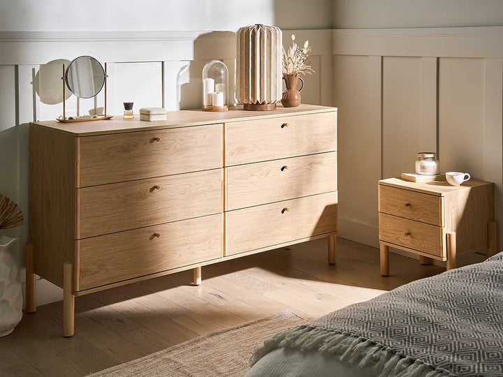 A light oak wood six-drawer dresser in a styled bedroom setting, accompanied by a matching nightstand and styled with home decor accessories.