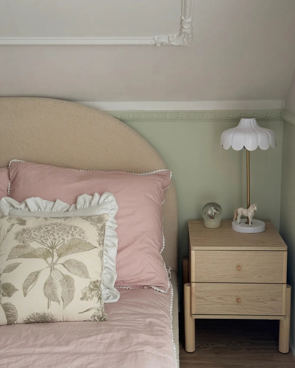 A styled bedroom corner featuring a beige upholstered bed headboard, pink bedding with decorative pillows, a light wood nightstand, and a white scalloped table lamp against a sage green wall.