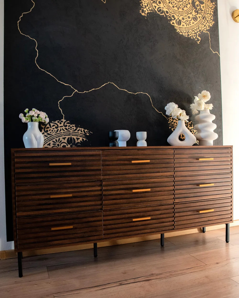 A mid-century modern wooden sideboard with a slatted drawer front and gold-tone handles, styled against a black wall with gold line art and white ceramic vases.