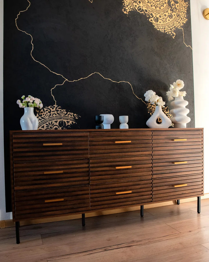 A mid-century modern wooden sideboard with a slatted drawer front and gold-tone handles, styled against a black wall with gold line art and white ceramic vases.